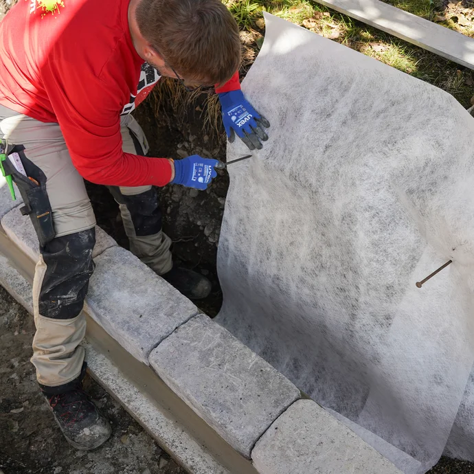 Heimwerker sichert das Drainagevlies mit langen Nägeln in der Baugrube im Garten.