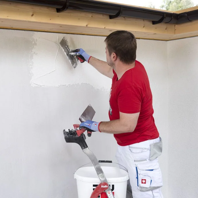 Mann steht auf Leiter und trägt Rillenputz von oben nach unten mit einer Glättkelle an der Wand auf.