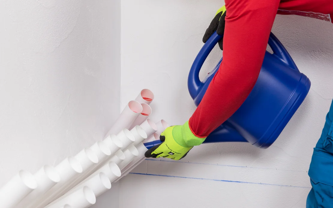 Person mit Schutzhandschuhen bef&uuml;llt mit einer blauen Gie&szlig;kanne Injektionstrichter in einer Wand zur Horizontalabdichtung.
