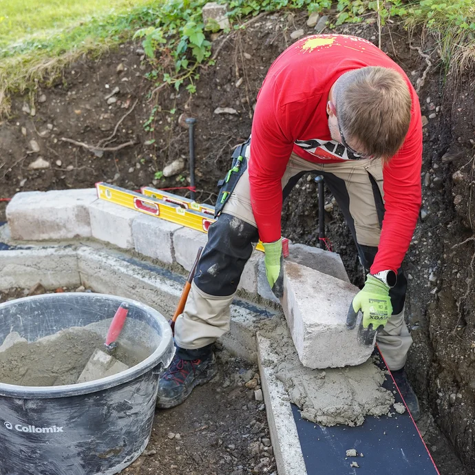 Eine Person setzt einen Mauerstein in einen frischen Mörtel auf eine Fundamentreihe. Eine Wasserwaage und ein Mörtelkübel mit angerührtem Mörtel und Kelle liegen daneben.