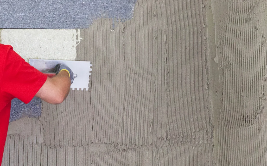 Person tr&auml;gt Kleber mit Zahnspachtel auf Wand auf, f&uuml;r Fliesenverlegung vorbereitet.