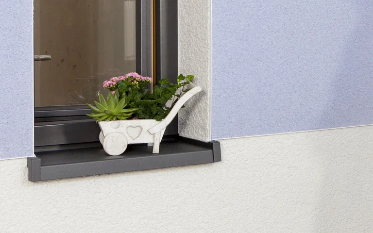 Dekorativ verputzte Fassade mit blauer Kratzputzfläche und Blumen am Fensterbrett.
