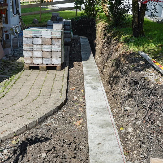 Frisch ausgeschaltes Streifenfundament entlang eines Gartenwegs, daneben gestapelte Pflastersteine und ein ausgehobener Erdstreifen.