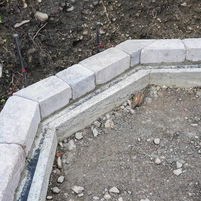 Das Bild zeigt eine frisch gesetzte erste Steinreihe einer Gartenmauer, die im Mörtelbett ausgerichtet wurde. Die Steine sind sauber platziert und liegen zum Trocknen bereit. Im Hintergrund sind Erdreich und Richtschnüre zur Ausrichtung zu sehen.