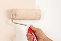Hand streicht mit einer Farbrolle hellrosa Farbe auf weiße Wand für effiziente Renovierung.