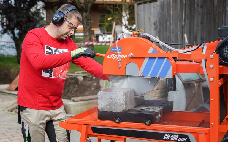 Heimwerker sägt Natursteine und schneidet Betonsteine im Garten mit der Norton Clipper Steinsäge, um eine Gartenmauer zu bauen. Er trägt Gehörschutz, Schutzbrille und Arbeitshandschuhe.
