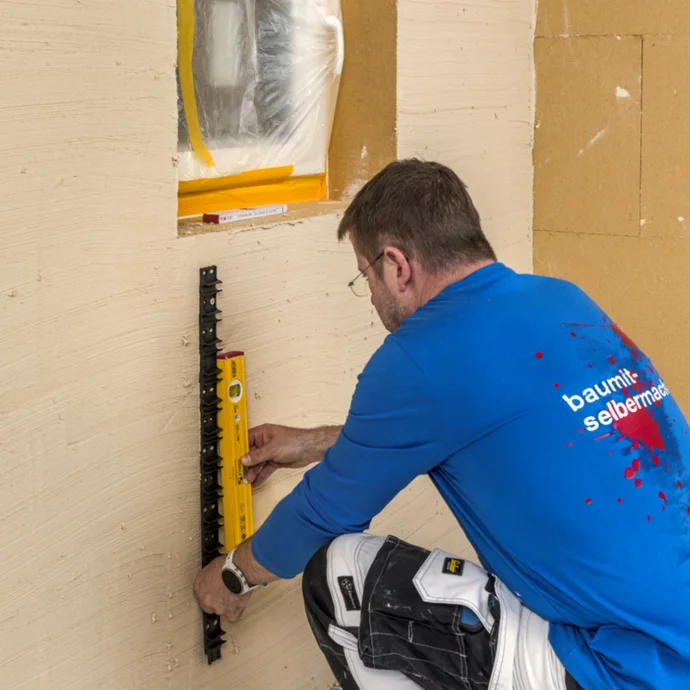 Mann misst Wand mit Wasserwaage, um Fensterrahmen pr&auml;zise einzubauen, vorbereitet mit Baumaterialien von Baumit.