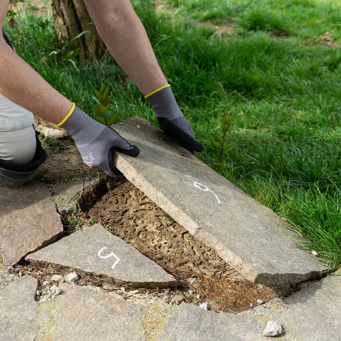 Handwerker hebt eine lose Natursteinplatte mit Handschuhen vom Boden ab.