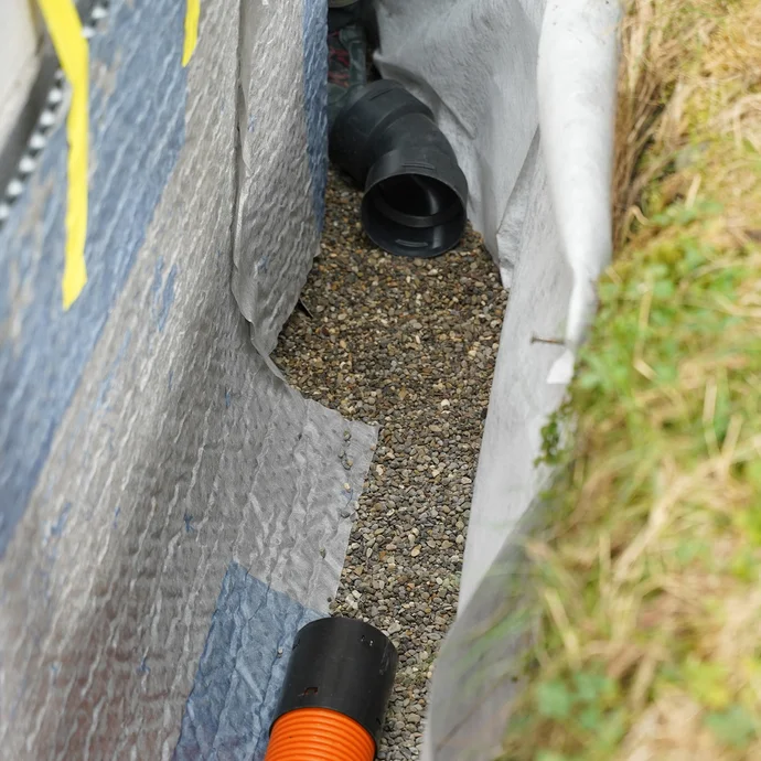 Detailaufnahme: schwarzes Winkelstück eines Drainagerohrs auf Filterkies. Auf der linken Seite ist die Drainagematte und auf der rechten Seite ist das eingebaute Drainagevlies erkennbar.