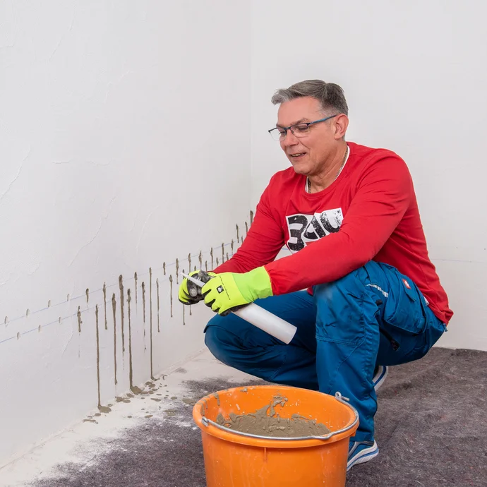 Handwerker bereitet einen weiteren Injektionstrichter vor. Die Einf&uuml;llspitze wird mit einem Cuttermesser gek&uuml;rzt. In einem Eimer ist der bereits angemischter Betonspachtel.