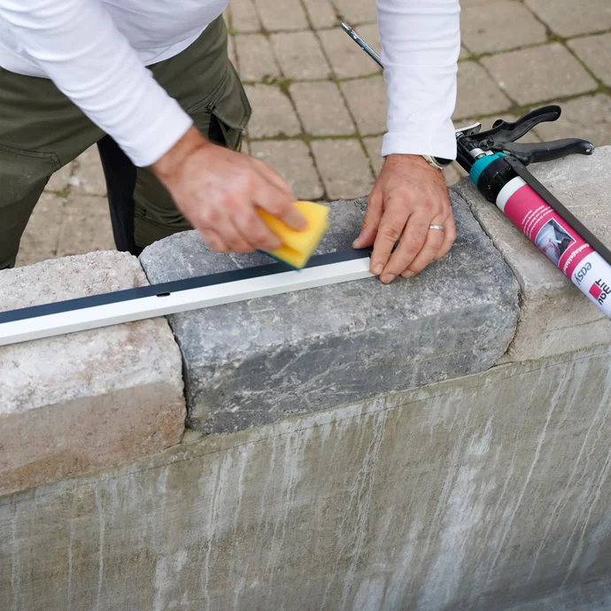 Handwerker reinigt auf einer Mauer die Klebeseite eines Abschlussprofils mit einem Schwamm. Eine Kartuschenpistole mit easy Montagekleber EVO liegt daneben.