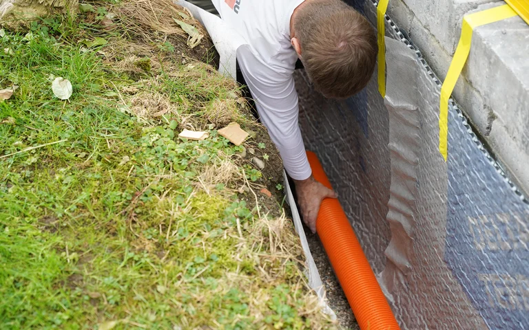 Handwerker verlegt ein Drainagerohr auf dem Filterkies an der Gartenmauer.